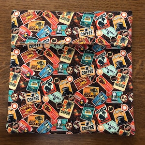 May include: A brown fabric lunch bag with a repeating pattern of vintage coffee advertisements. The advertisements feature various coffee blends, coffee shops, and coffee beans. The bag has a flap that folds over and closes with a velcro strap.