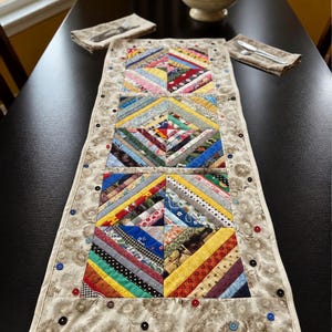 May include: A rectangular table runner featuring three square quilt blocks made of multicolored fabric strips in a diagonal pattern. The runner has a beige floral border embellished with small, colorful buttons sewn along the edges.
