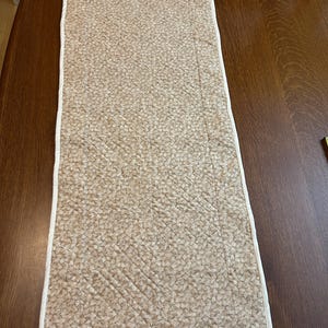 May include: A rectangular quilted table runner featuring a light tan fabric with a dense, repeating white floral pattern. The runner is finished with a clean white fabric binding along all four edges and displays visible diagonal stitching.