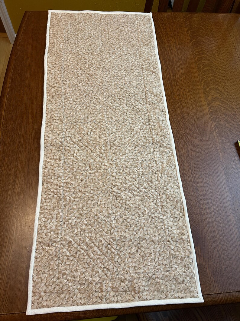 May include: A rectangular quilted table runner featuring a light tan fabric with a dense, repeating white floral pattern. The runner is finished with a clean white fabric binding along all four edges and displays visible diagonal stitching.