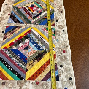 May include: A rectangular fabric table runner featuring a patchwork design of multi-colored fabric strips arranged in geometric patterns. The runner is bordered by a cream-colored floral fabric adorned with various buttons. A yellow measuring tape shows a length of approximately 40 inches.