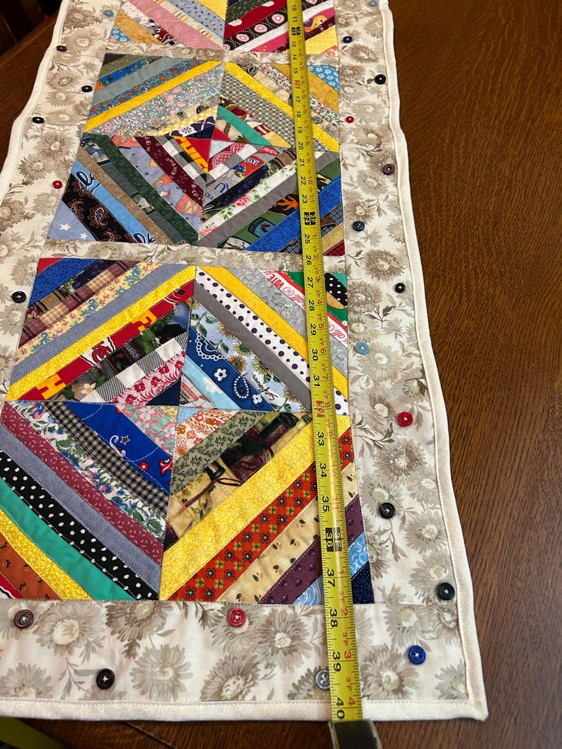 May include: A rectangular fabric table runner featuring a patchwork design of multi-colored fabric strips arranged in geometric patterns. The runner is bordered by a cream-colored floral fabric adorned with various buttons. A yellow measuring tape shows a length of approximately 40 inches.