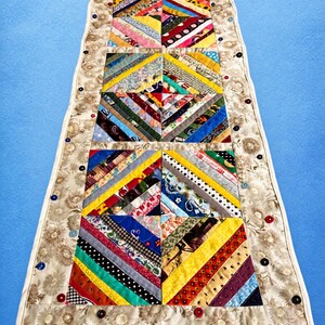May include: A rectangular fabric table runner featuring a central patchwork design of multi-colored diagonal fabric strips arranged in square blocks. The runner is bordered by a beige floral fabric embellished with small, colorful buttons.