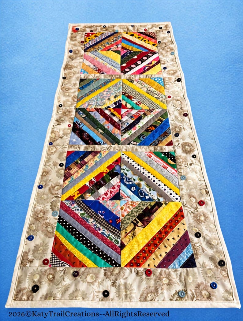 May include: A rectangular fabric table runner featuring a central patchwork design of multi-colored diagonal fabric strips arranged in square blocks. The runner is bordered by a beige floral fabric embellished with small, colorful buttons.