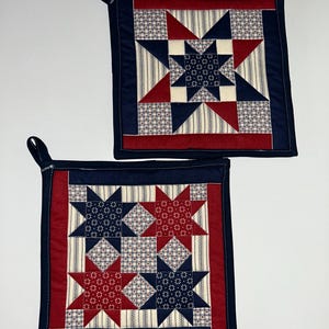 May include: Two square quilted pot holders with a patriotic star pattern. Each pot holder features a central star design in navy blue and red, surrounded by a cream-colored background with a small print. The borders are navy blue and red.