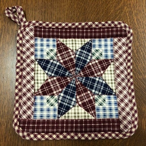 May include: A red, white, and blue plaid quilted hot pad with a starburst design. The hot pad has a loop handle for hanging.