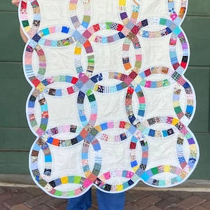 May include: A handmade quilt featuring a double wedding ring pattern. The quilt has a white background with colorful rings made of various patterned fabric squares. The edges are scalloped and outlined in white. A unique, handcrafted textile.