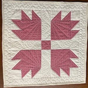 May include: A square, handmade quilt featuring a central design of four stylized bear paws in a deep rose color with a small floral pattern. The paws are set against a cream-colored background with intricate quilted patterns. The quilt has a cream-colored border.