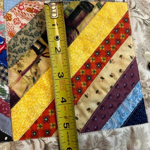 May include: A patchwork quilt featuring diagonal strips of various patterned fabrics in yellow, red, blue, and floral prints. A yellow measuring tape is placed vertically across the fabric, showing a length of approximately 6.5 inches.