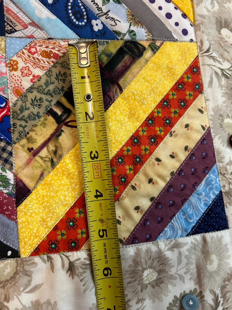 May include: A patchwork quilt featuring diagonal strips of various patterned fabrics in yellow, red, blue, and floral prints. A yellow measuring tape is placed vertically across the fabric, showing a length of approximately 6.5 inches.