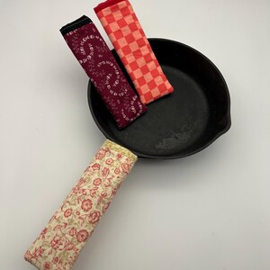 May include: A black cast iron skillet with three fabric handle covers. Two covers are on the skillet, one with a red and white checkered pattern and the other with a burgundy floral design. The third cover has a cream floral pattern.