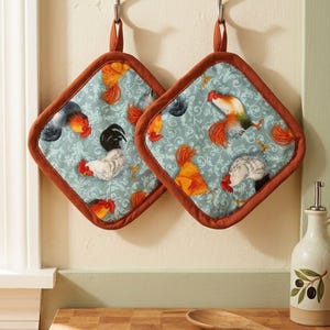 May include: Two diamond-shaped pot holders featuring a light blue fabric with a white damask pattern and a repeating rooster print in shades of red, orange, and black. Each pot holder has a solid rust-colored fabric trim and a matching hanging loop.