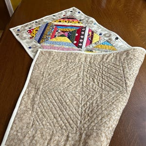 May include: A reversible quilted table mat featuring a colorful patchwork side with various fabric patterns and a solid beige side with a quilted diamond and line texture. The border is finished with white binding and small decorative buttons.