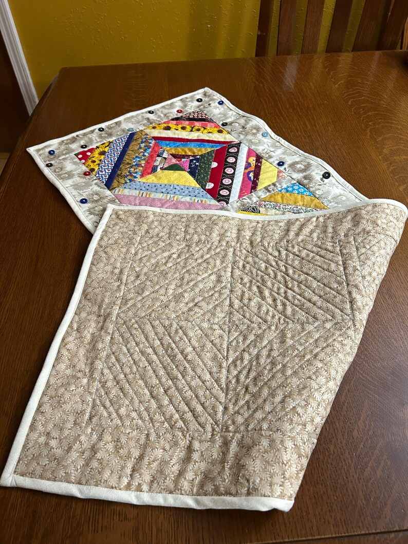 May include: A reversible quilted table mat featuring a colorful patchwork side with various fabric patterns and a solid beige side with a quilted diamond and line texture. The border is finished with white binding and small decorative buttons.