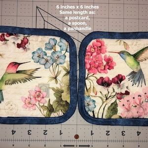 May include: A pair of 6 inch by 6 inch pot holders with a blue trim. The pot holders feature a floral pattern with hummingbirds and a vintage postcard design. The text on the pot holders reads "6 inches x 6 inches Same length as: a postcard, a spoon, a panhandle"
