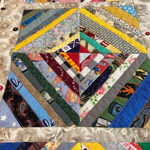 May include: A rectangular fabric table runner featuring a multi-colored string quilt pattern with diagonal strips of various patterned cotton fabrics. The border is a beige floral print fabric accented with small, round, multi-colored buttons.