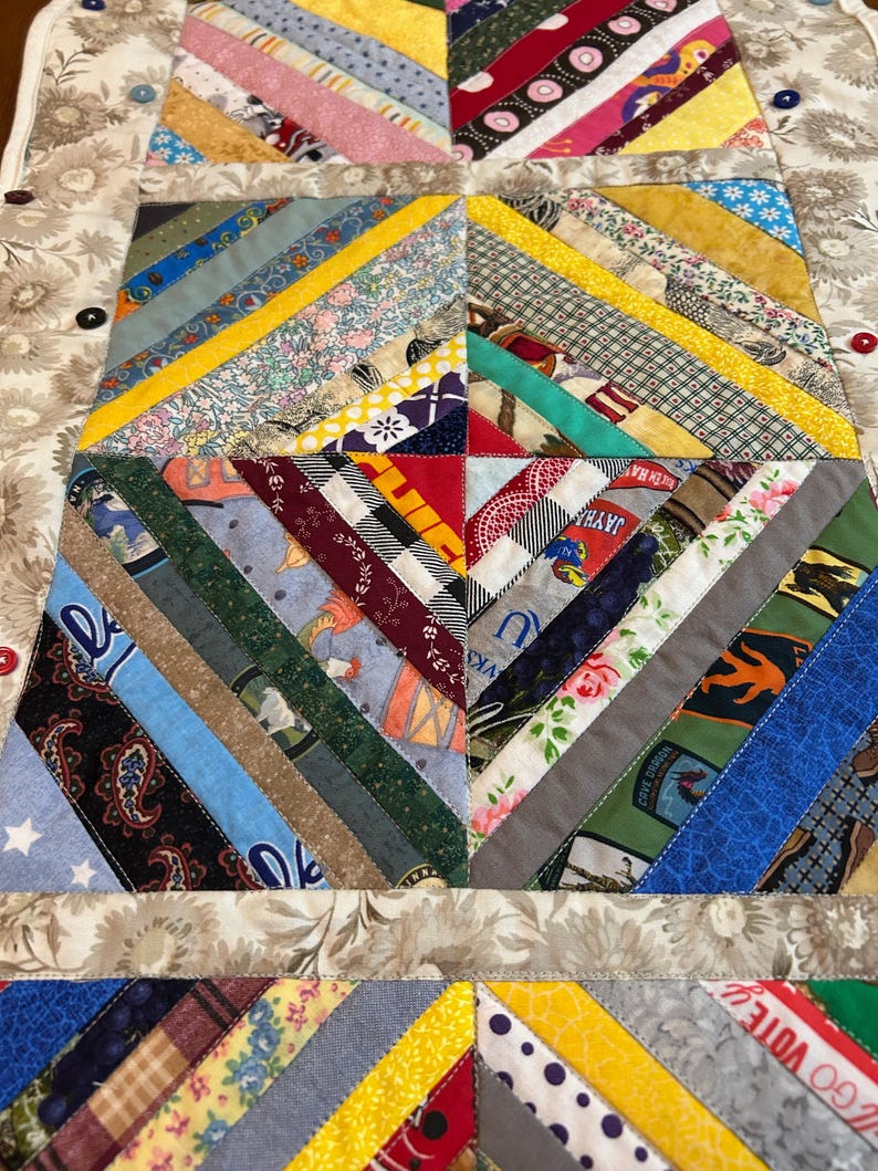 May include: A rectangular fabric table runner featuring a multi-colored string quilt pattern with diagonal strips of various patterned cotton fabrics. The border is a beige floral print fabric accented with small, round, multi-colored buttons.