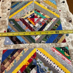May include: A rectangular fabric table runner featuring a multi-colored string quilt pattern with various printed cotton strips. A yellow measuring tape across the center shows a width of approximately 17 inches. The border is a beige floral fabric.