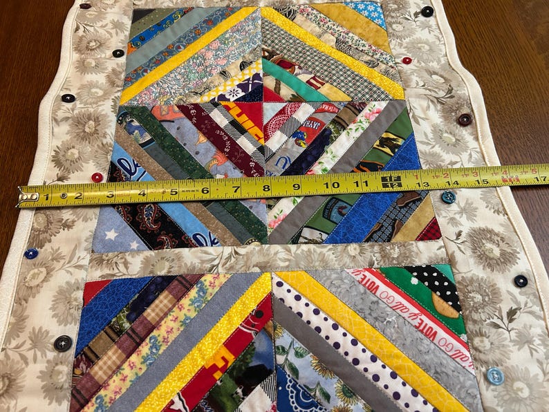 May include: A rectangular fabric table runner featuring a multi-colored string quilt pattern with various printed cotton strips. A yellow measuring tape across the center shows a width of approximately 17 inches. The border is a beige floral fabric.