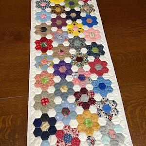 May include: A handmade table runner featuring a floral design. The runner is made of white fabric with a quilted pattern. The flowers are made of various colorful fabrics, including red, blue, yellow, and green. The runner is approximately 40 inches long.