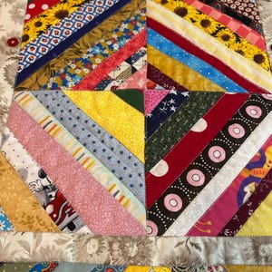 May include: A square fabric quilt block featuring a diagonal string-pieced pattern with various colorful cotton prints including sunflowers, polka dots, and stars. The border is a beige floral fabric accented with small red, blue, and black buttons.
