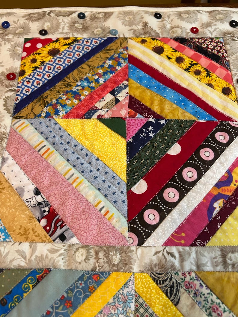 May include: A square fabric quilt block featuring a diagonal string-pieced pattern with various colorful cotton prints including sunflowers, polka dots, and stars. The border is a beige floral fabric accented with small red, blue, and black buttons.