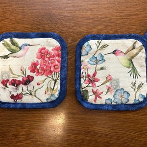 May include: Two square pot holders with a blue border and hanging loop. Each features a hummingbird and floral design in pink, red, and blue. The background has a vintage, aged paper appearance.