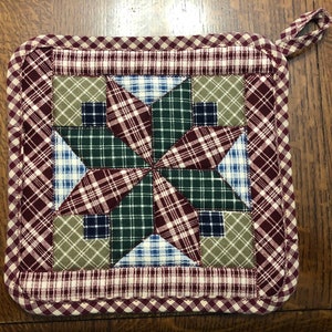 May include: Square quilted potholder with a starburst design. The center features a star pattern in green, burgundy, blue, and tan plaid fabrics. The border is a burgundy and white checkered pattern, with a loop for hanging.
