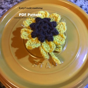 May include: A crocheted sunflower with a brown centre and yellow petals. The sunflower is sitting on a yellow ceramic pot lid. The text "PDF Pattern" and "Copyright © 2016 KatyTrailCreations" is visible on the lid.