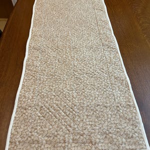 May include: A rectangular quilted table runner featuring a tan and beige fabric with a dense, repeating leaf pattern. The runner is finished with a clean, solid white fabric binding along all four edges, showcasing visible diagonal stitching.