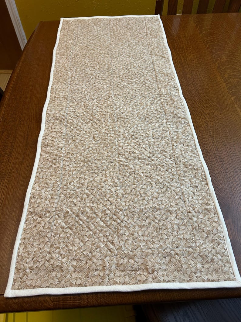 May include: A rectangular quilted table runner featuring a tan and beige fabric with a dense, repeating leaf pattern. The runner is finished with a clean, solid white fabric binding along all four edges, showcasing visible diagonal stitching.