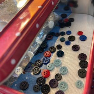 May include: A red metal tin with a white polka dot lid, partially open to reveal a collection of assorted plastic buttons in various colors including black, blue, red, and light blue, scattered inside on a patterned paper liner.