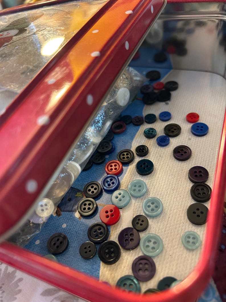 May include: A red metal tin with a white polka dot lid, partially open to reveal a collection of assorted plastic buttons in various colors including black, blue, red, and light blue, scattered inside on a patterned paper liner.
