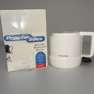 Peut inclure: Une bouilloire électrique sifflante blanche Proctor Silex avec une poignée et un bouton d'alimentation noir. La boîte à côté porte le logo Proctor Silex et des informations sur le produit. La bouilloire contient 1 litre.
