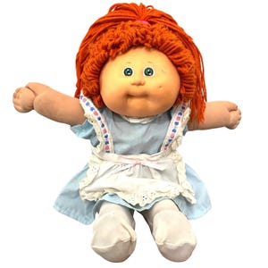 Puede incluir: Una muñeca Cabbage Patch Kid vintage con cabello de hilo naranja y ojos verdes. La muñeca lleva un vestido azul claro con un delantal blanco y medias blancas. Los brazos de la muñeca están extendidos.
