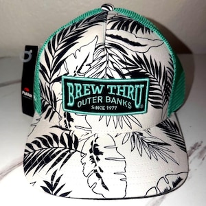 May include: A white and black patterned baseball cap with a green mesh back. The cap features a patch that reads "BREW THRU OUTER BANKS SINCE 1977". The hat has a curved brim and a tag attached.