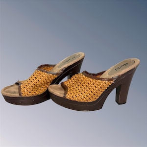 May include: A pair of Mudd brand platform mules featuring a woven tan upper, an open toe, and a dark brown block heel. The footbed is light tan with the Mudd logo printed in black. The design includes a thick platform sole and brown trim.