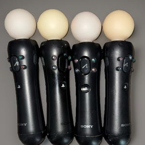May include: Four black PlayStation Move controllers with white and off-white spheres on top. Each controller has multiple buttons and the Sony logo. The controllers are arranged vertically against a gray background.