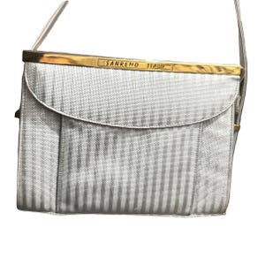 May include: A silver-colored, rectangular handbag with vertical stripes. The bag has a gold-colored frame and a matching shoulder strap. The text "SANREMO ITALY" is displayed on the frame.