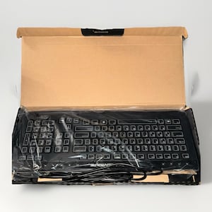 May include: A black computer keyboard inside its cardboard box. The keyboard is rectangular with numerous keys and a black cable. The box is open, revealing the keyboard wrapped in plastic. The brand name is visible on the keyboard.