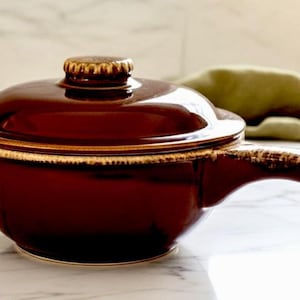 May include: A dark brown ceramic soup bowl with a lid. The bowl has a handle and a gold-colored trim around the rim. The lid has a small, decorative knob. The bowl is on a white marble surface.
