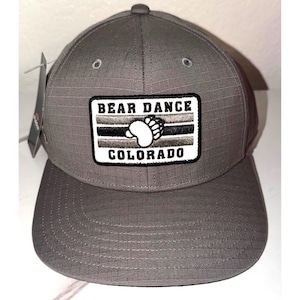 May include: Gray baseball cap with a structured design. The cap features a rectangular patch with the words "BEAR DANCE COLORADO" and a bear paw graphic. The cap has a curved brim and a button on top.