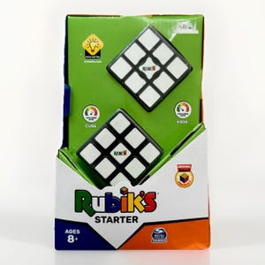 Puede incluir: Un rompecabezas de cubo Rubik's Starter en su embalaje original. La caja es blanca, verde y naranja, con el logotipo de Rubik's y el texto "Starter" en un lugar destacado. El rompecabezas es blanco y negro, con el logotipo de Rubik's en cada cuadrado. A partir de 8 años.