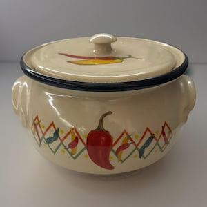 Peut inclure: Un plat à casserole en céramique crème avec un couvercle, bord noir et deux poignées. Le plat est décoré d'un piment rouge et d'autres poivrons colorés. Le couvercle a un petit bouton et un motif de piment.