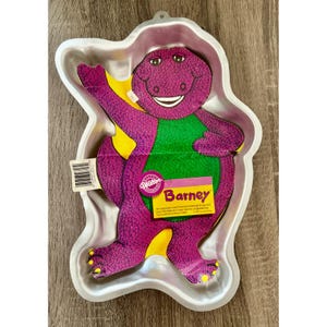 May include: A silver-colored metal cake pan in the shape of Barney the dinosaur. The purple dinosaur has a green belly and yellow accents. The pan includes a paper tag with the name "Barney" and the Wilton logo.