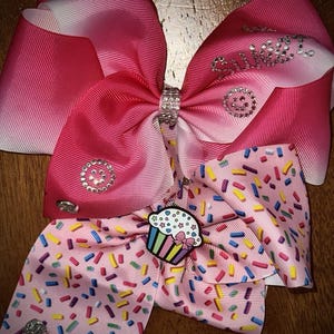 May include: Two large, pink hair bows. The top bow is ombre, fading from white to pink. The bottom bow is light pink with colorful sprinkles and a cupcake charm. Both bows have rhinestone accents and a rhinestone-studded center.