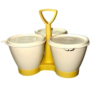 Puede incluir: Un especiero Tupperware vintage en crema y amarillo. El especiero tiene tres recipientes de color crema con tapas blancas, unidos por un asa amarilla. La base también es amarilla. Los recipientes están diseñados para contener condimentos.