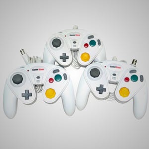 Pode incluir: Três controladores GameStop brancos com botões de cores variadas e um direcional. Cada controlador tem um joystick cinza, um botão vermelho, verde e amarelo, e um direcional em forma de cruz. Os controladores são conectados por cabos brancos.
