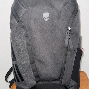 May include: A gray backpack with a textured fabric. It features a silver alien head logo, a black zipper, and black straps. The backpack has a padded back and multiple compartments for storage.