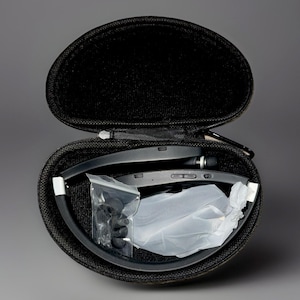 May include: Black, foldable Bluetooth headset with extra ear tips and accessories, inside a black, zippered, oval carrying case. The headset is folded and has buttons on the side. The case is open, revealing the contents.